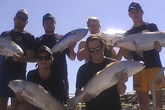 Six people holding large fish, standing outside under a clear sky.